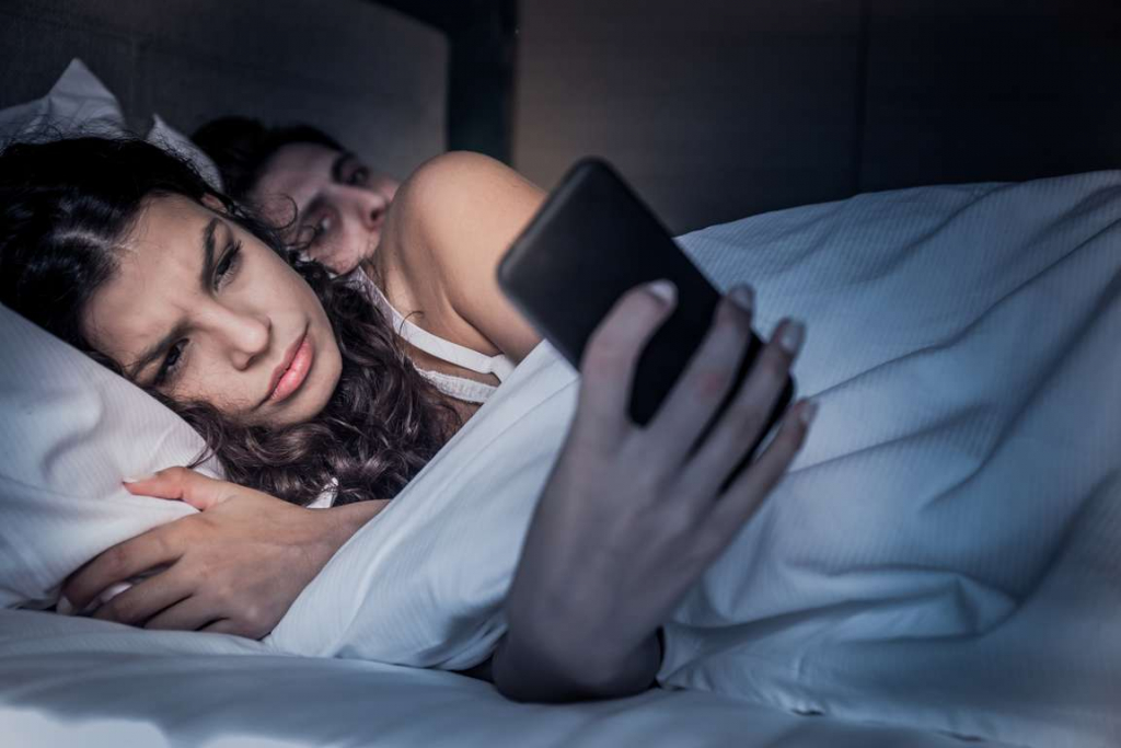 Digital Infidelity reading secret messages in bed at night while partner looks over shoulder